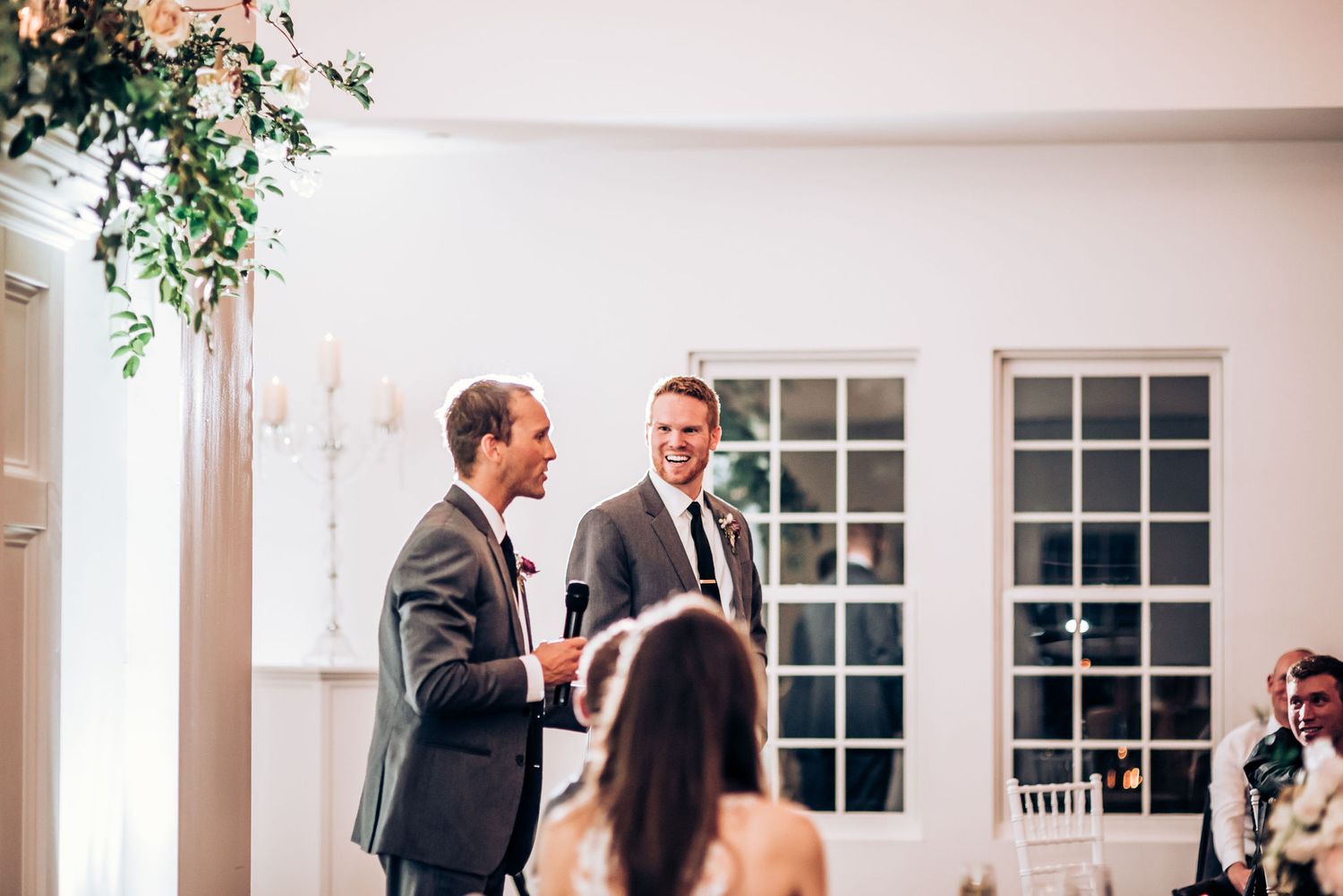 Two people in formal wear speaking to wedding guests at an elegant white-walled venue.