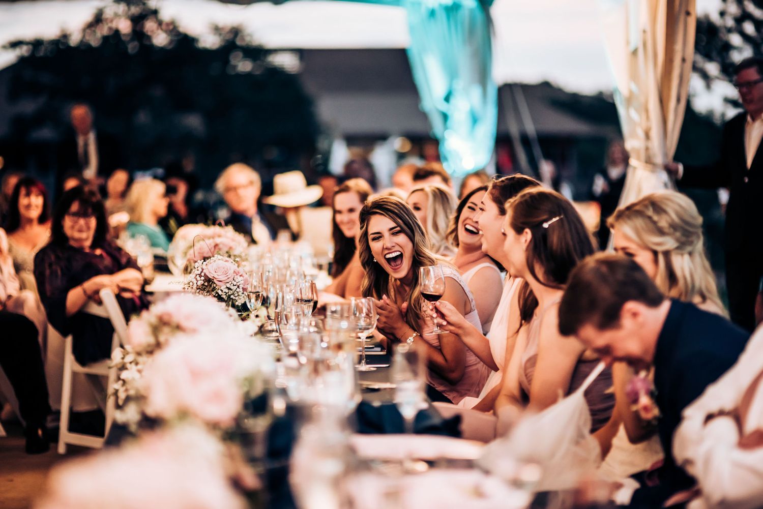 Wedding guests laughing and enjoying speeches during outdoor reception.