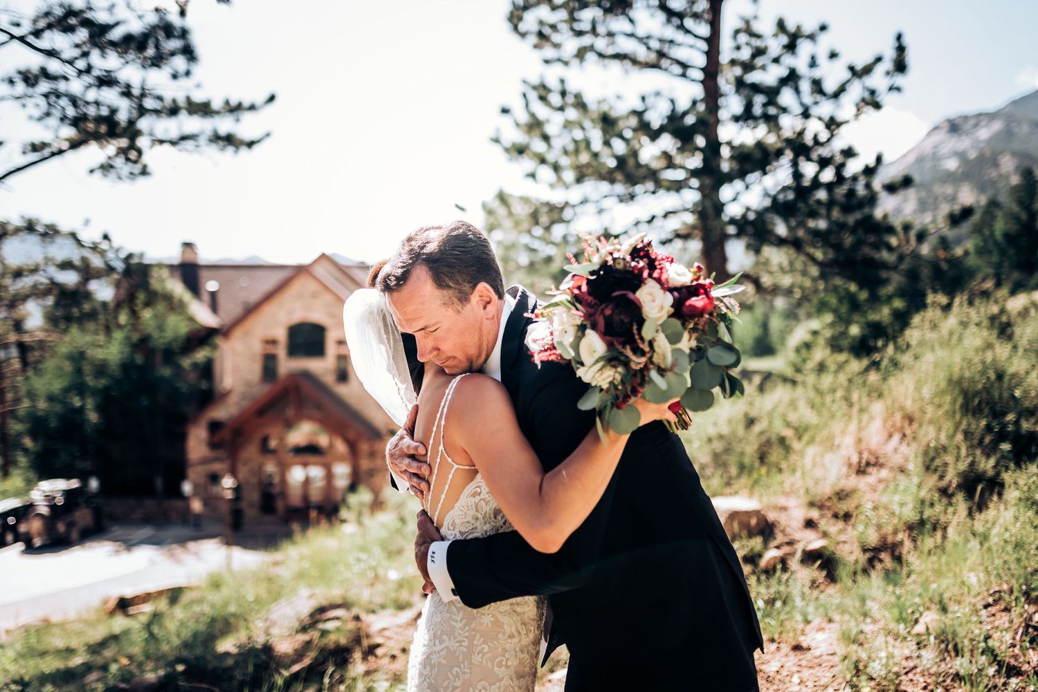 Two people embrace during an outdoor mountain wedding ceremony with a rustic cabin in the background.