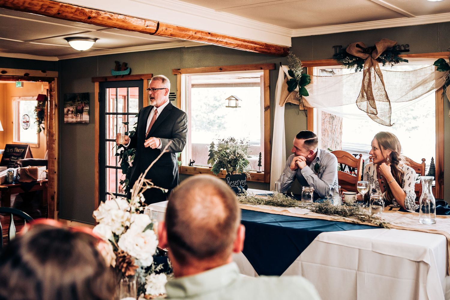 A speaker addresses wedding guests during a reception in a warmly lit rustic venue.