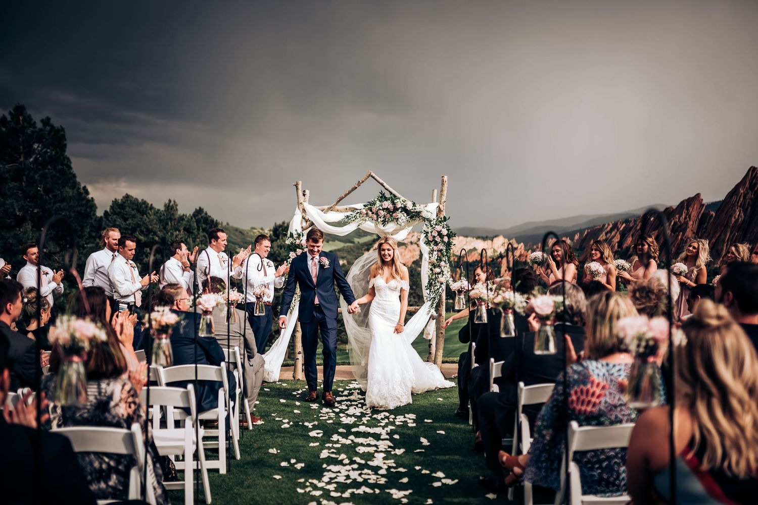 Scenic mountain wedding ceremony with white flower arch and rows of white chairs on lawn.