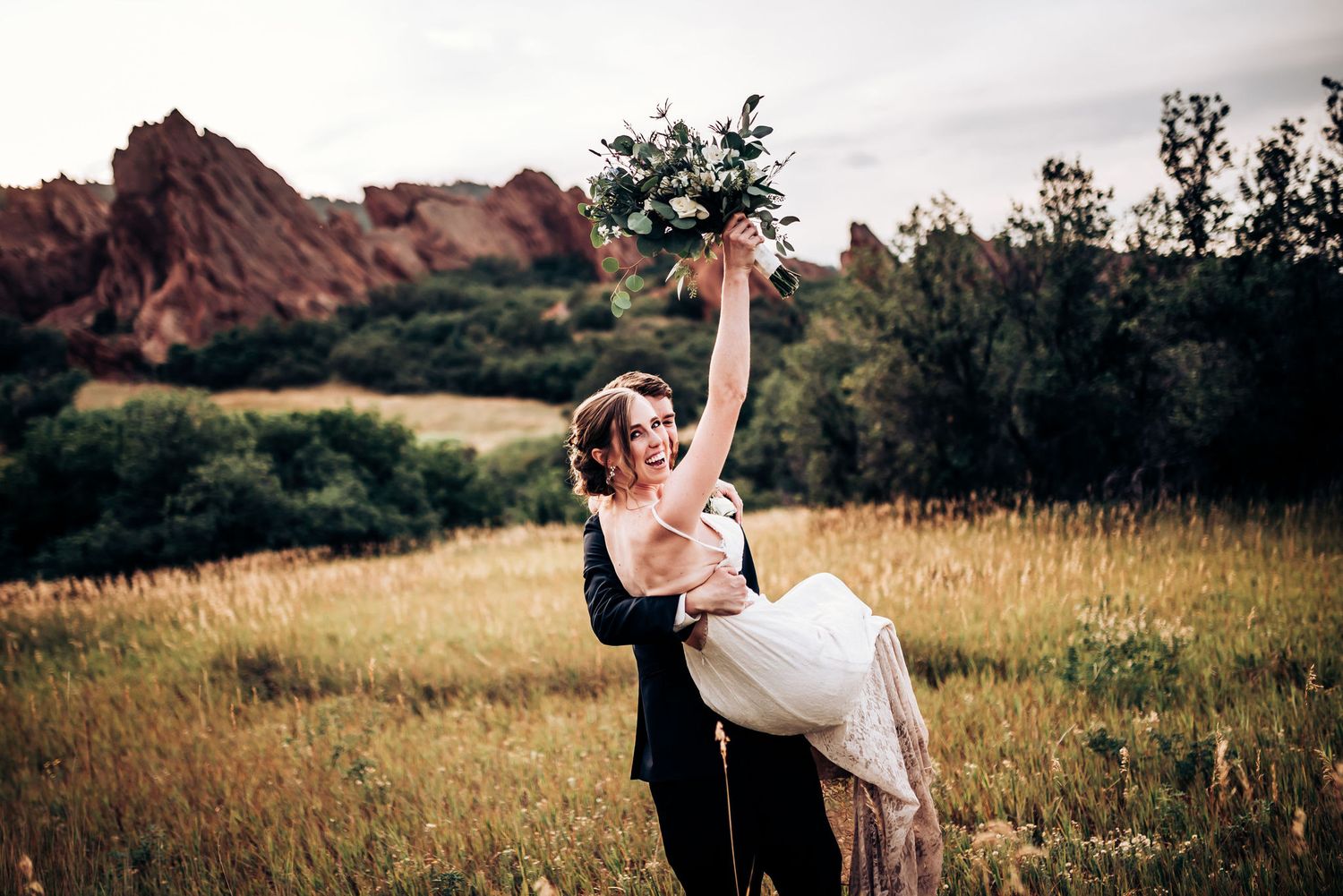 Groom lifts bride in white dress against backdrop of red rock formations.