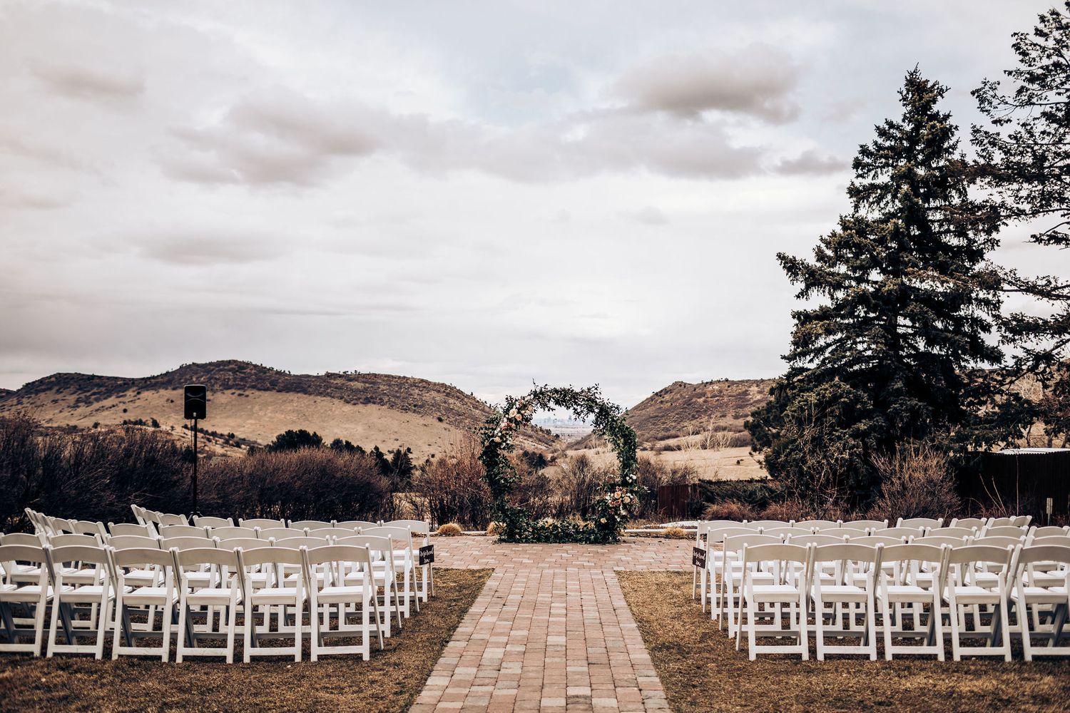 Outdoor wedding ceremony setup with white chairs and floral arch against mountain backdrop.
