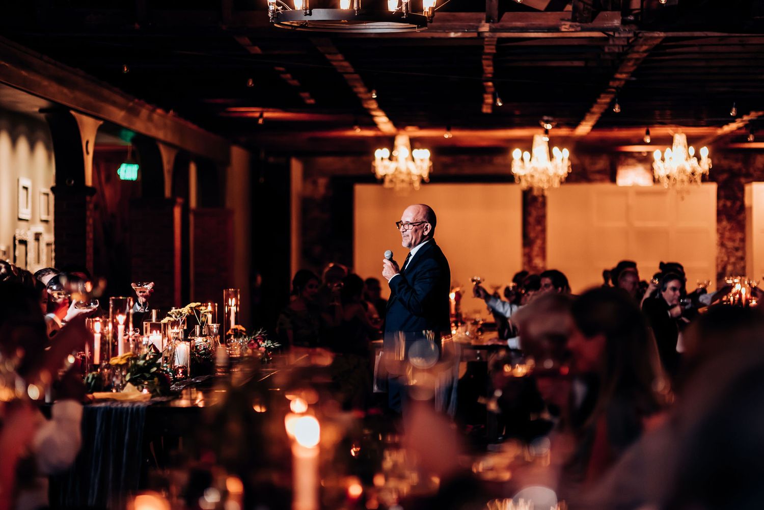 Speaker stands among dinner tables at an evening reception with warm lighting and chandeliers.