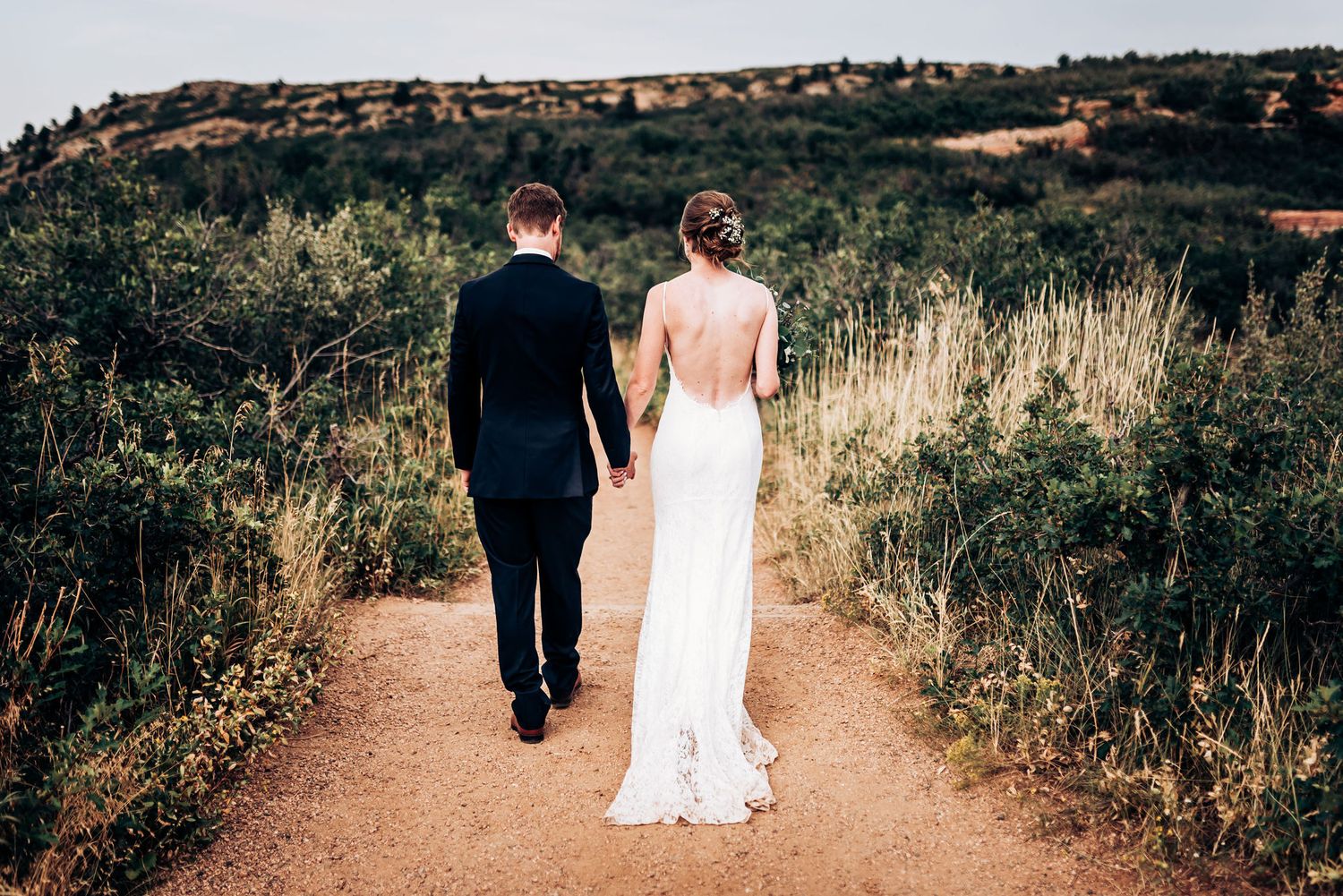 A couple walks hand in hand down a dirt path surrounded by natural landscape.