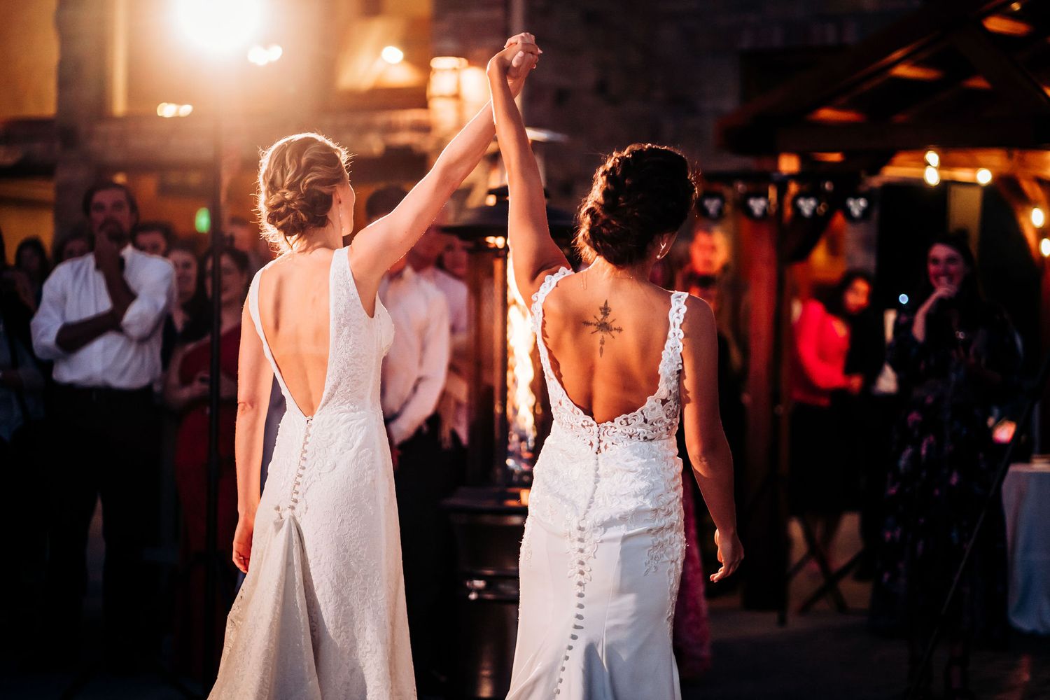 Two people in backless white dresses raise joined hands during a dance celebration.