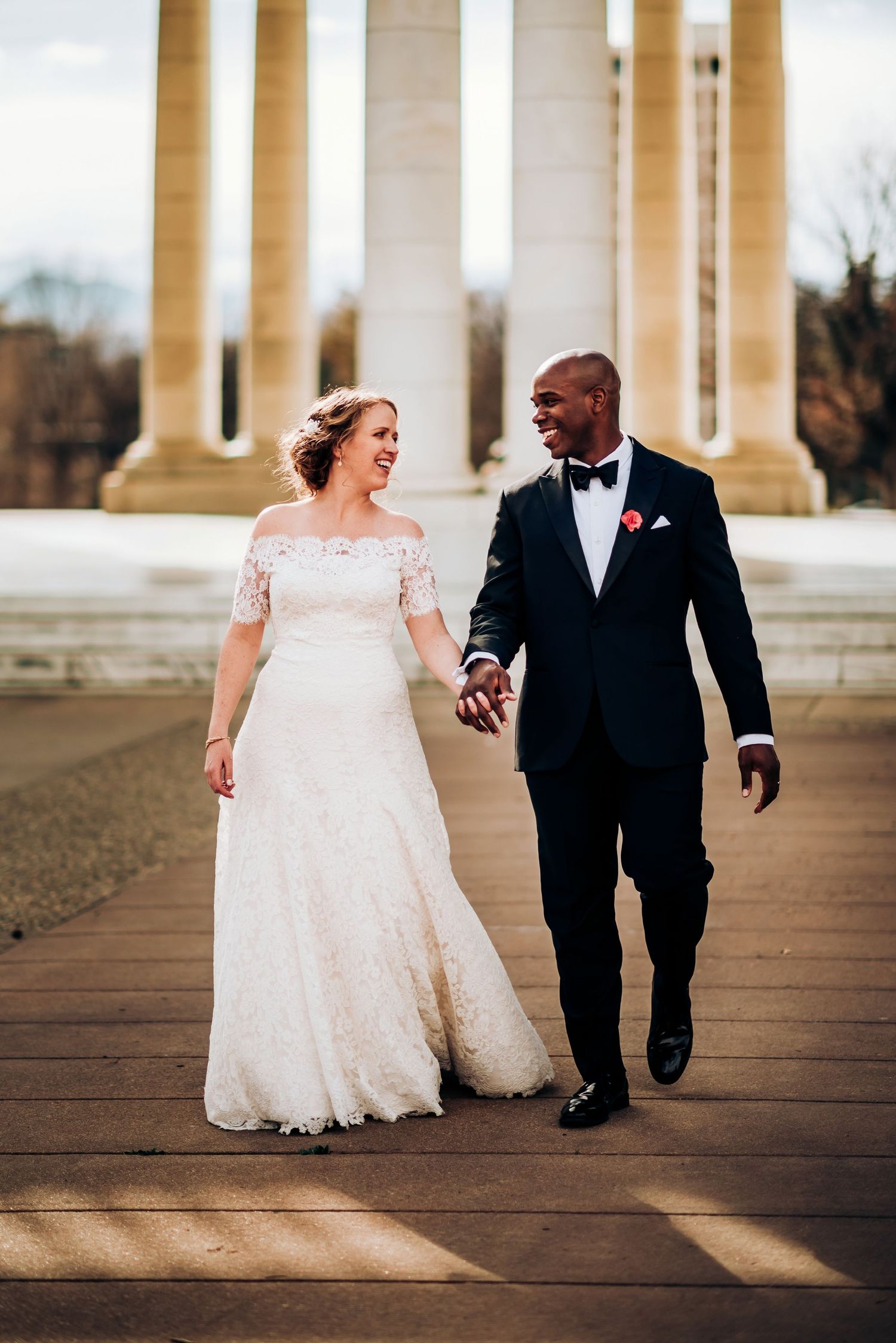 Newlyweds walk together between elegant white columns in formal wedding attire.