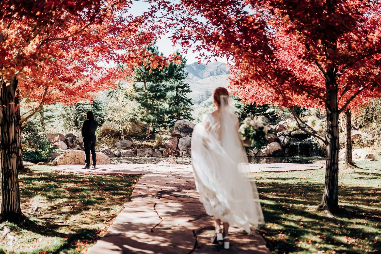 Wedding couple walks through autumn trees with vibrant red foliage.