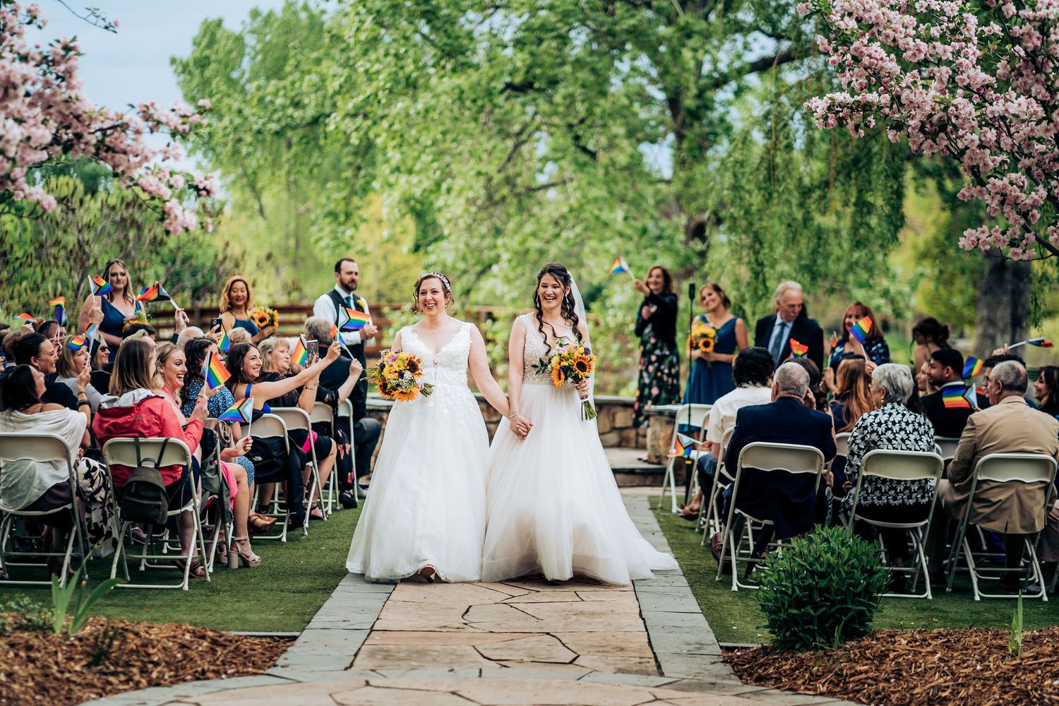 Two brides walk down the aisle together at an outdoor garden ceremony.