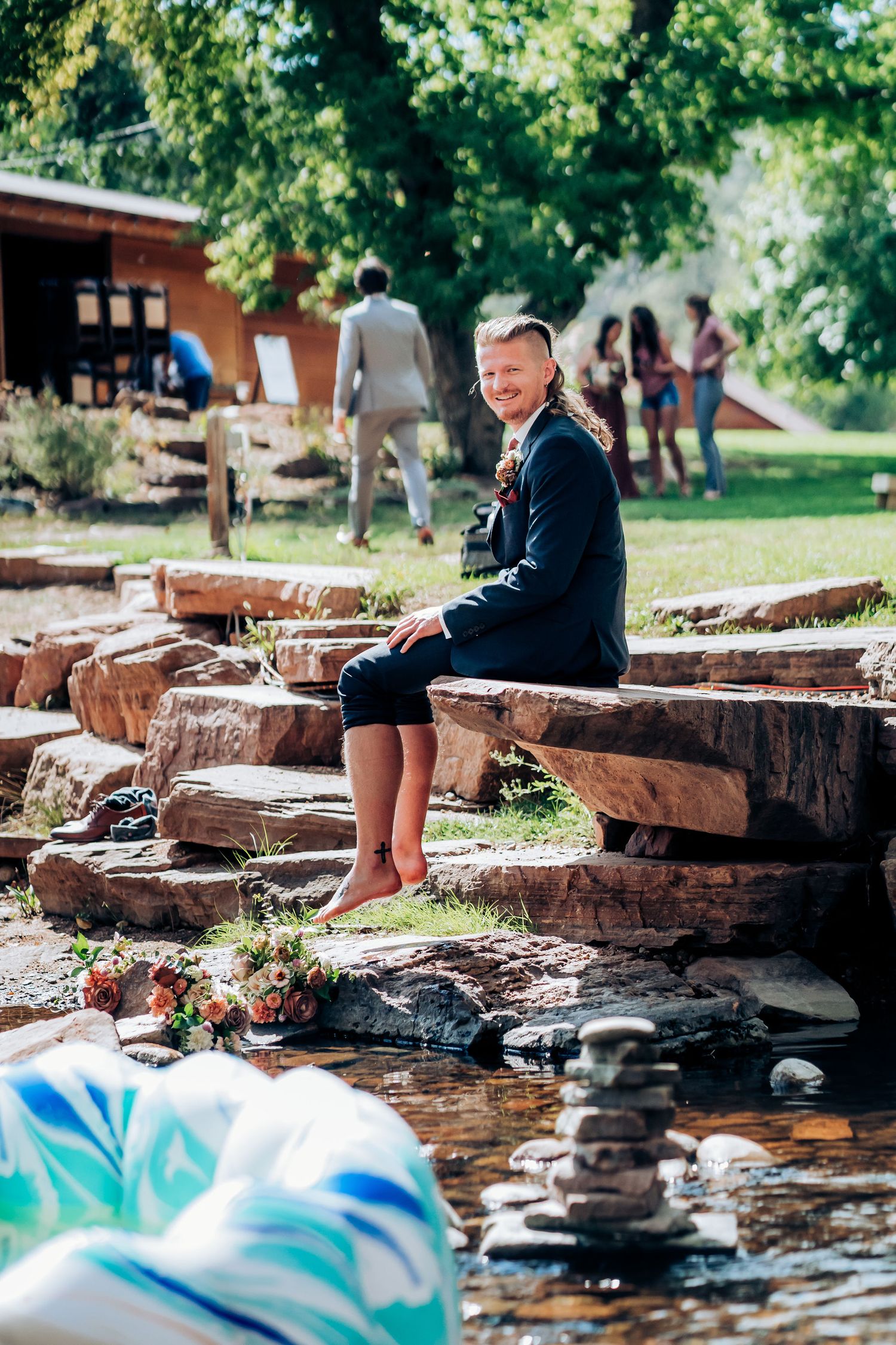 Wedding guest relaxes on stone ledge near water during outdoor celebration.