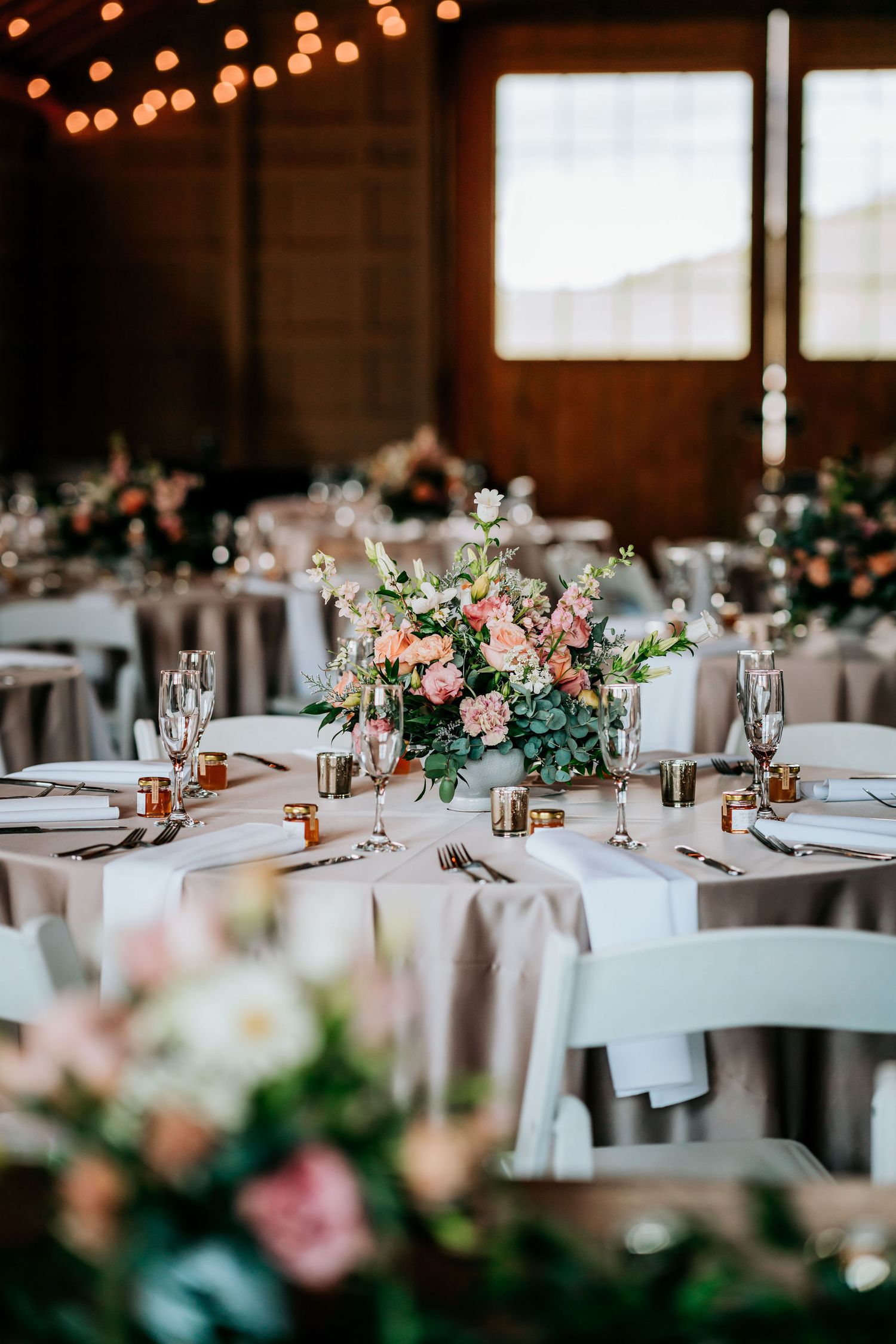 Wedding reception tables with pink floral centerpieces and white chairs in barn setting.