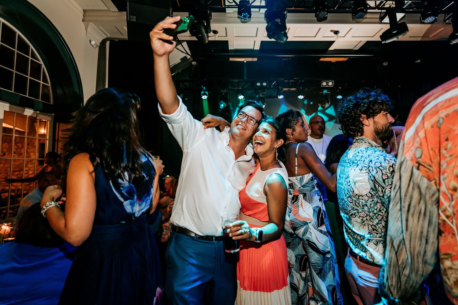 Celebration at indoor venue with guests dancing under blue lighting and arched windows.