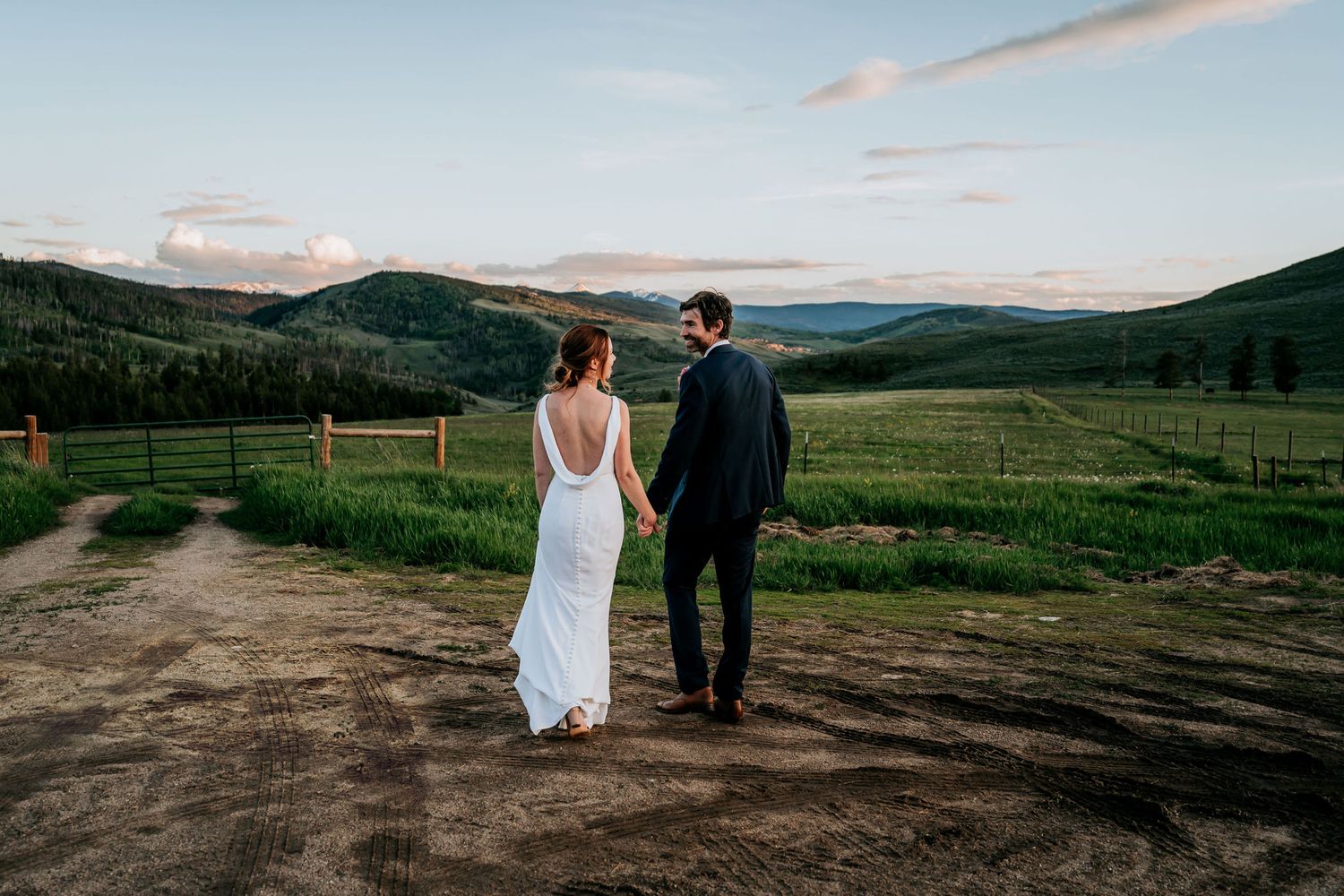 A couple walks hand in hand on a rural path surrounded by green mountain vistas.