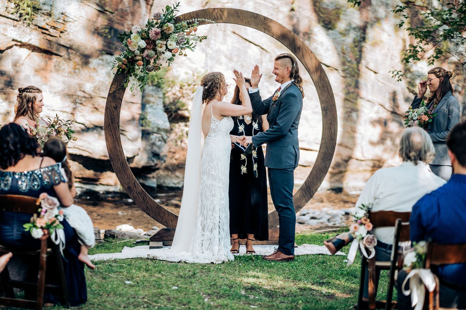 Wedding ceremony under a circular arch decorated with florals against rocky landscape.