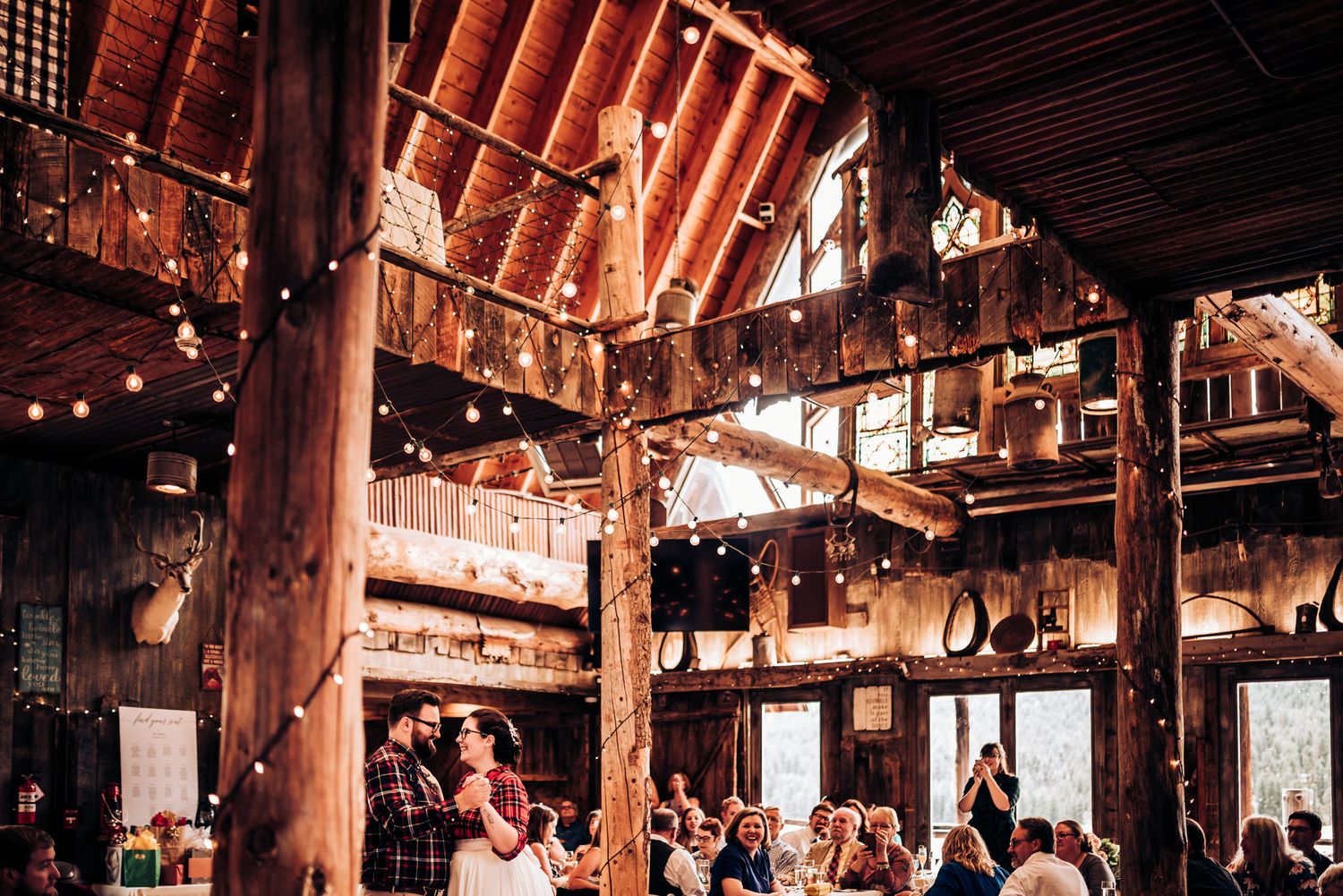 Interior of a rustic barn venue with exposed wooden beams and string lights during a wedding reception.