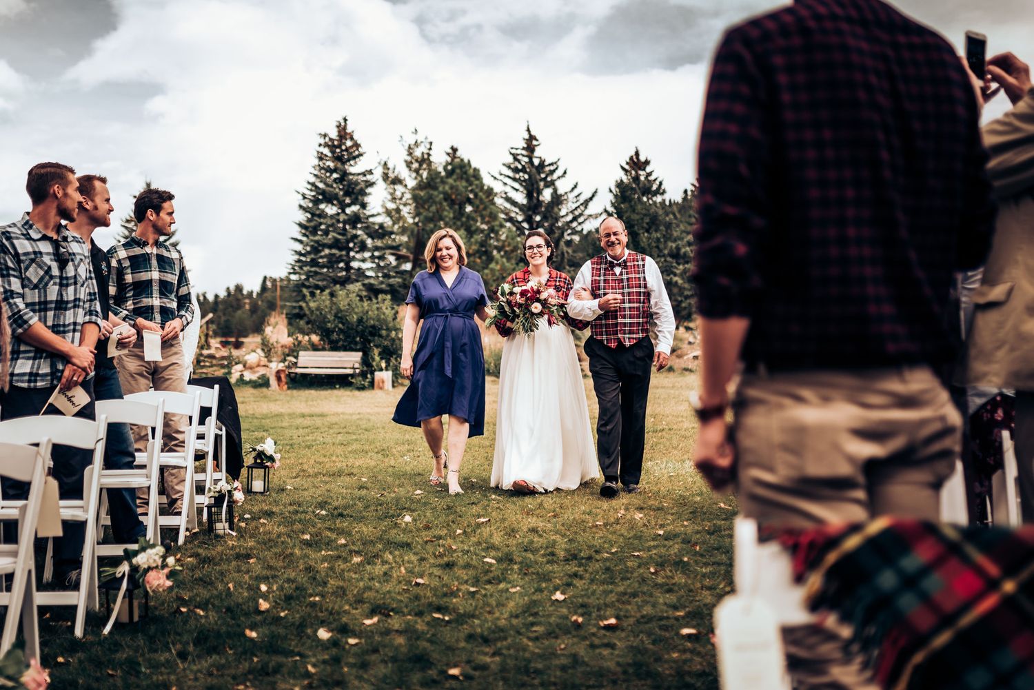 Outdoor wedding ceremony procession on grass lawn with guests seated in white chairs.
