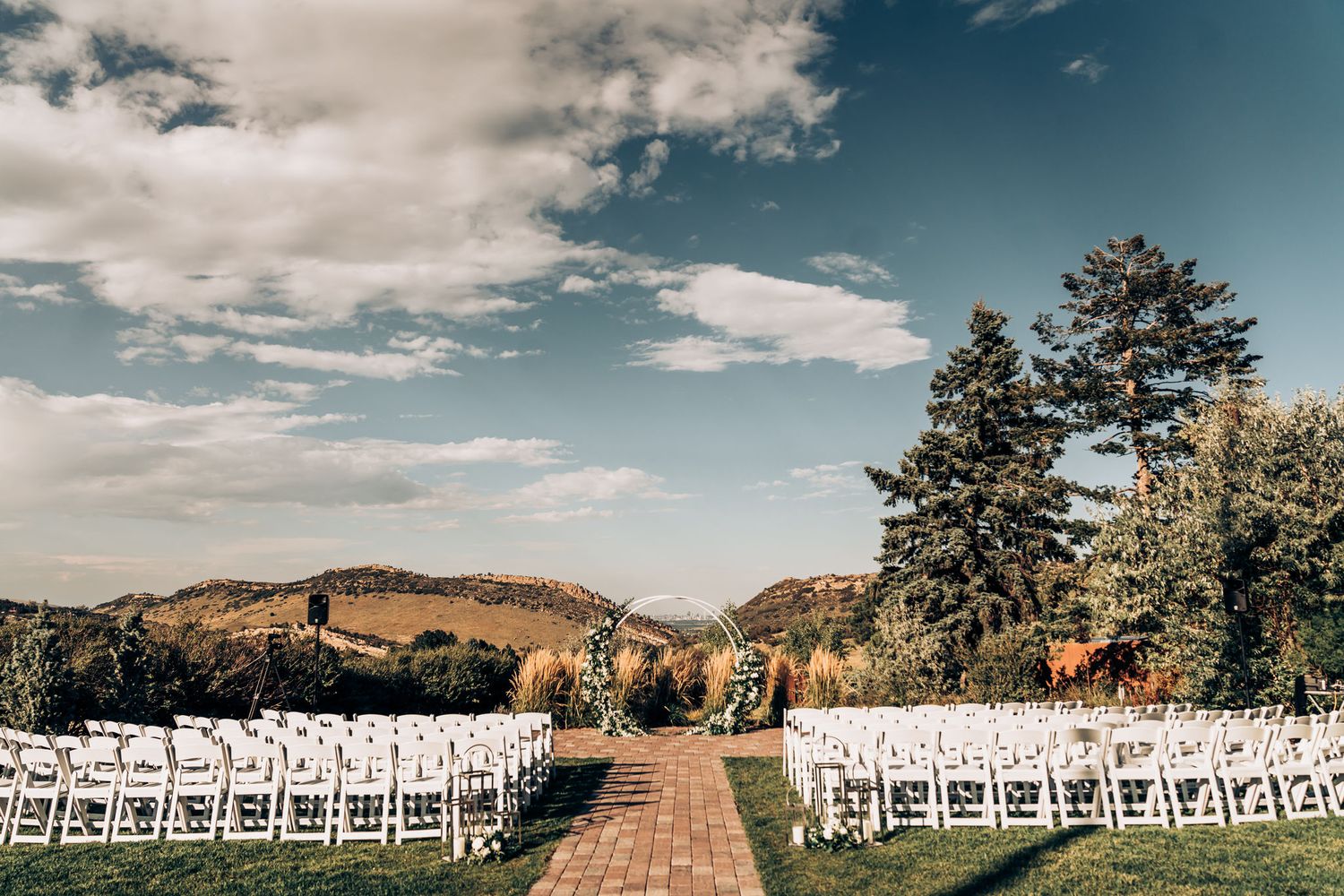 White chairs line both sides of an outdoor wedding aisle with mountain views and blue skies.