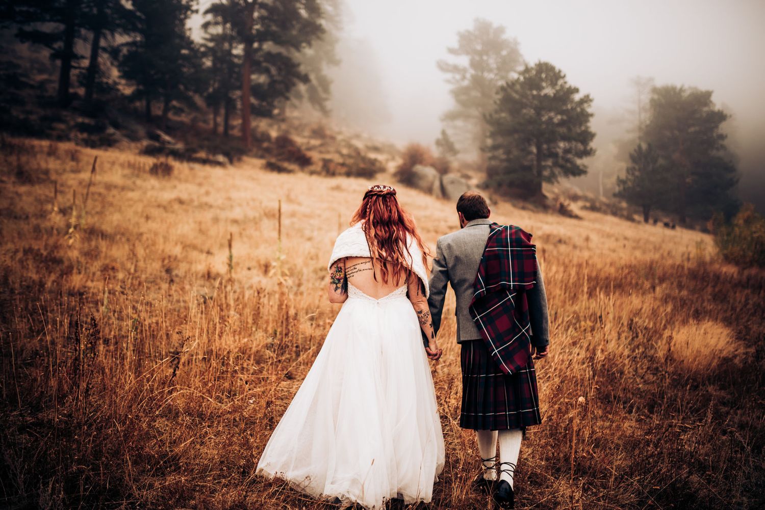 Couple walking hand in hand through golden mountain field with groom wearing traditional kilt.