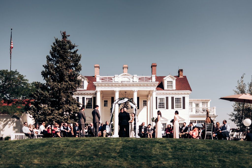 Large white colonial mansion hosting outdoor wedding with guests gathered on the lawn.