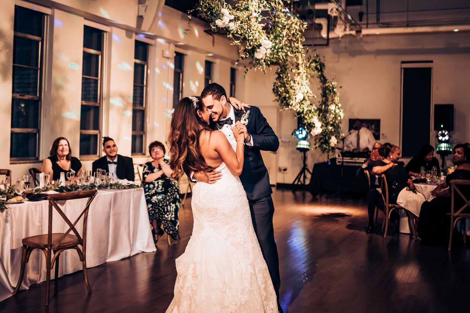 A couple embraces during their wedding dance beneath elegant floral arrangements and string lights.