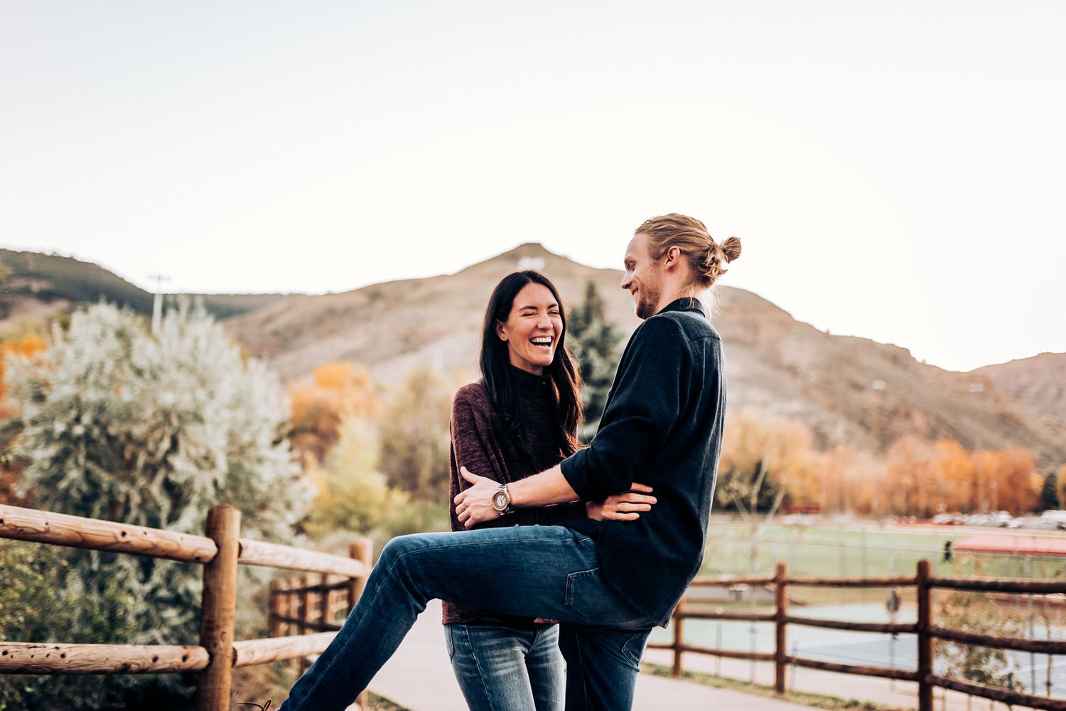 A couple sharing a playful moment on a wooden fence with autumn mountains in the background.