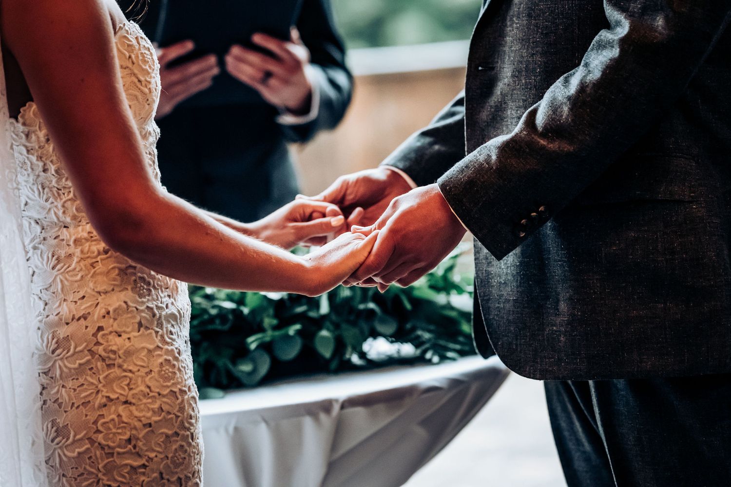 Close-up shot of bride and groom holding hands during their wedding ceremony with lace dress detail.