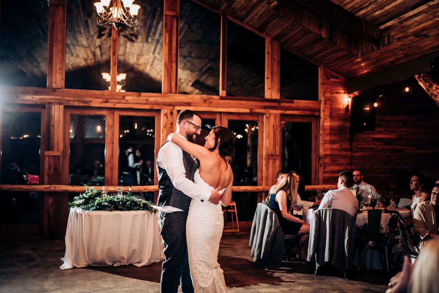 Newlyweds dance together in a rustic barn with large windows and elegant table settings.