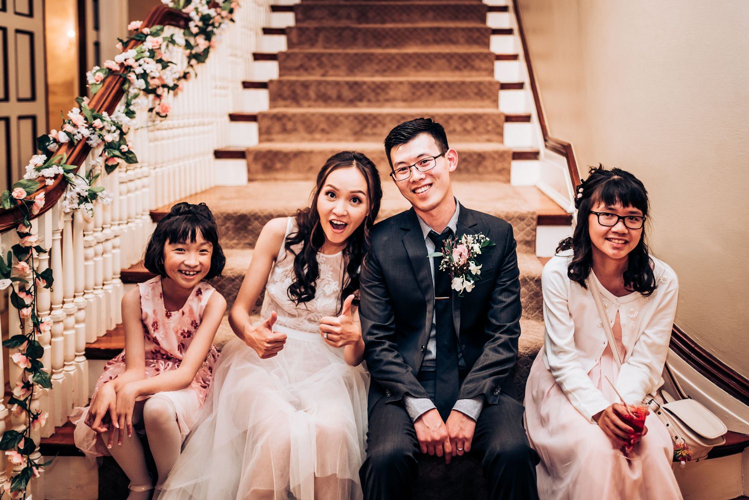 Wedding party sits together on staircase decorated with floral arrangements.