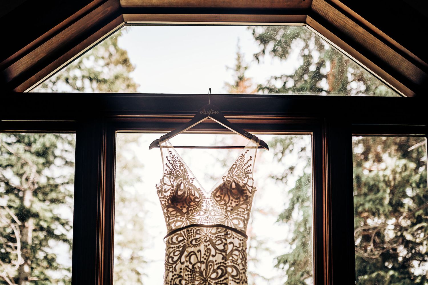 A detailed lace wedding dress hangs in front of a window with forest views.