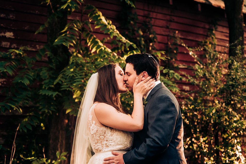 Romantic wedding scene with bride and groom sharing a kiss against a red brick wall with lush greenery.