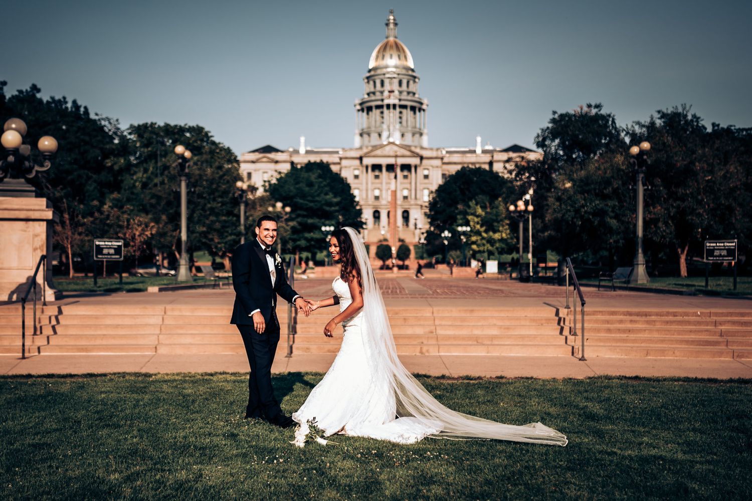 A wedding couple poses in front of a grand capitol building at dusk with a long flowing veil.