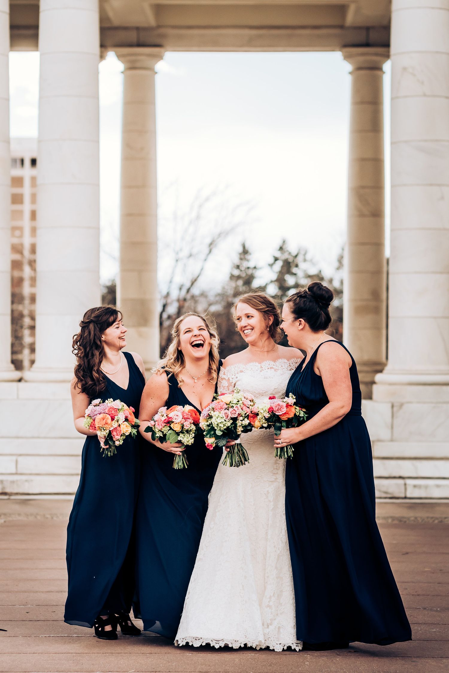 Candid moment of bridal party laughing together under elegant stone colonnade.