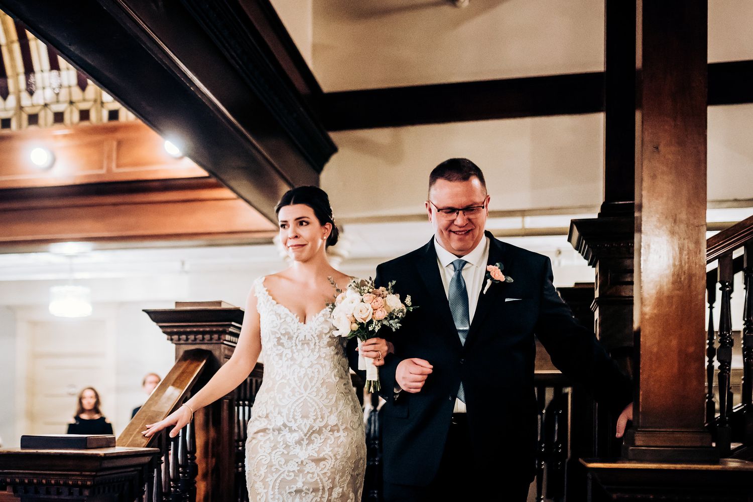 A couple descends a grand wooden staircase during a formal celebration.