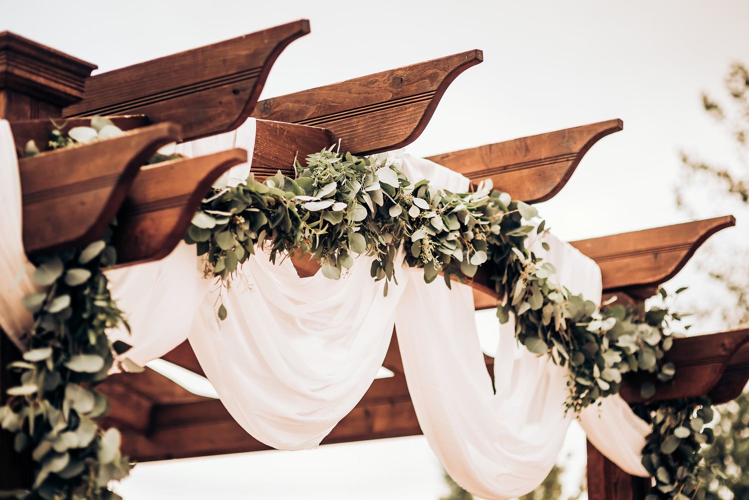 White draping and greenery decorate a wooden outdoor wedding ceremony arch.