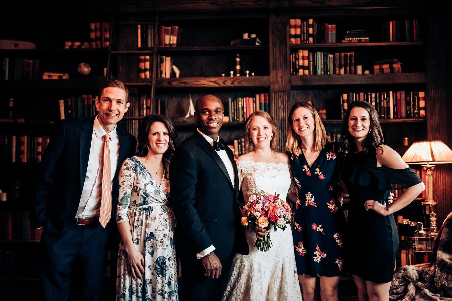 Wedding party poses together in library with bookshelves and warm lighting.