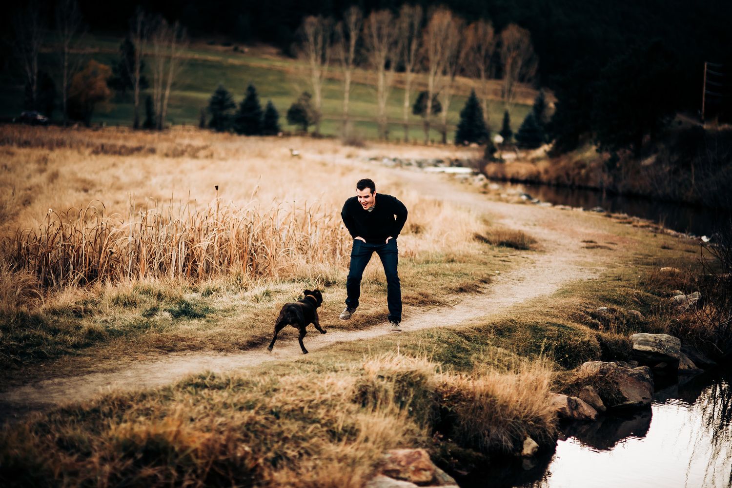 Person playing with dog on winding dirt path near water.