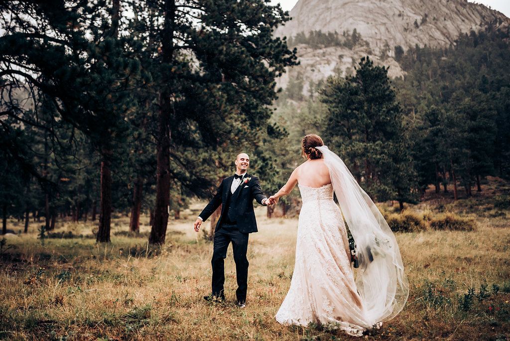 Wedding couple walks hand in hand through a scenic mountain meadow surrounded by pine trees.