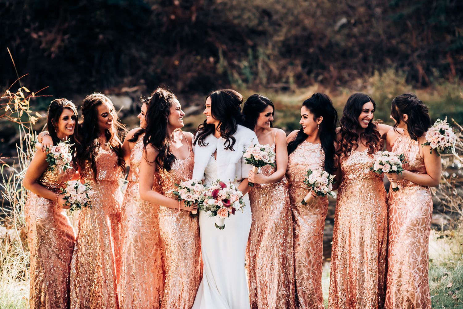 Wedding party poses outdoors with bridesmaids in sparkly rose gold dresses and bride in white.
