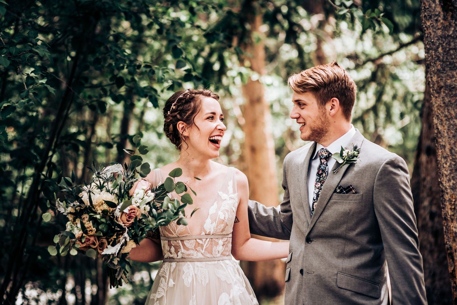 Candid laughter between bride and groom in wooded wedding setting with floral bouquet.