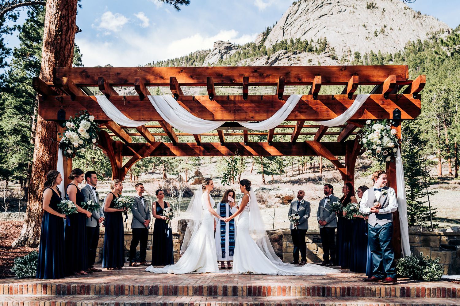 Wedding party assembled under decorated pergola against mountain landscape.