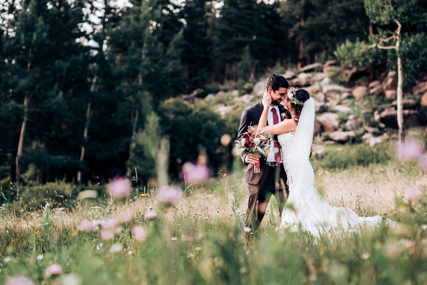 Newlyweds embrace in a sun-drenched mountain meadow with wildflowers and evergreen trees in the background.
