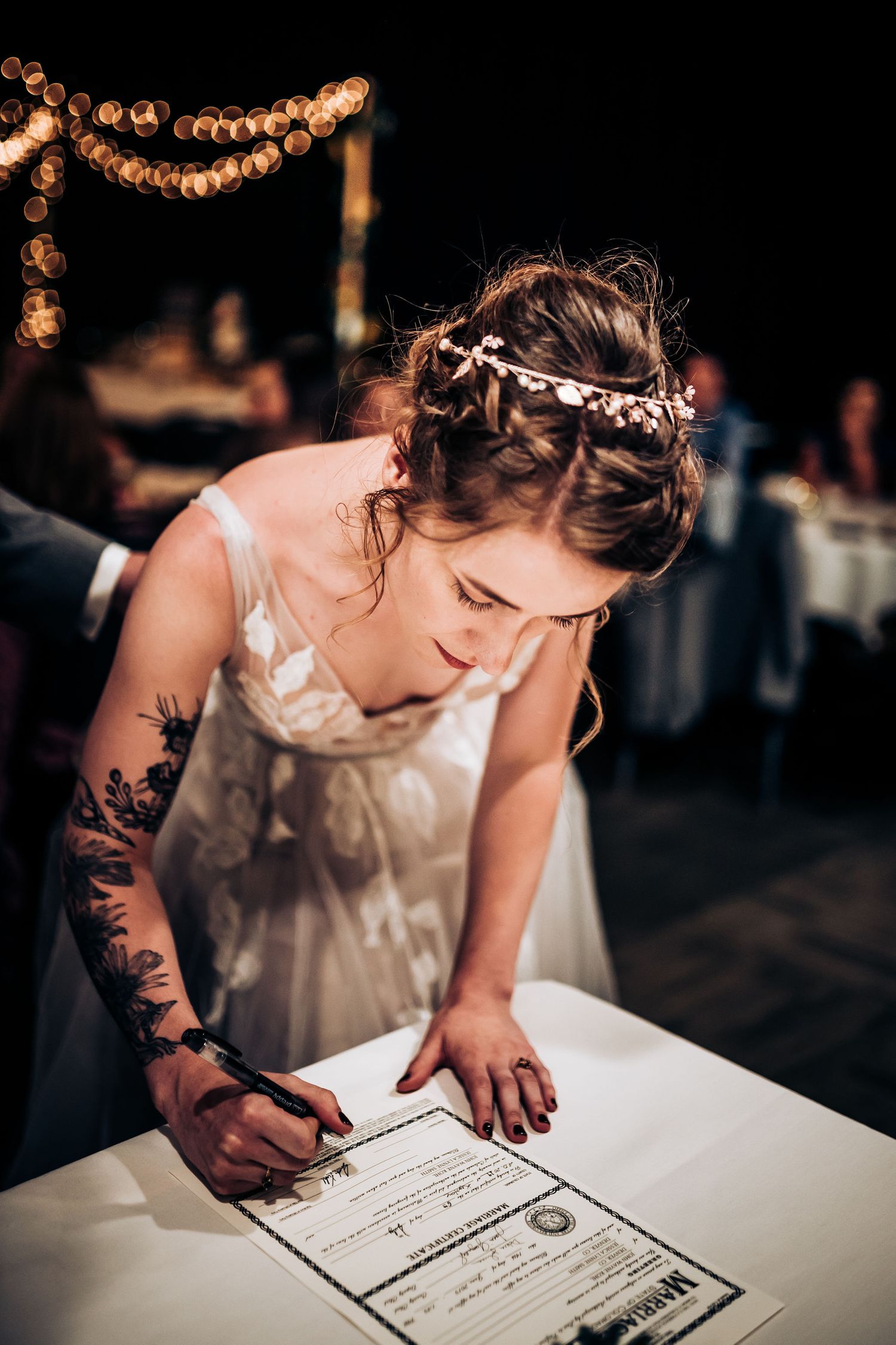 Bride in white dress with floral crown signs marriage certificate under string lights at evening reception.