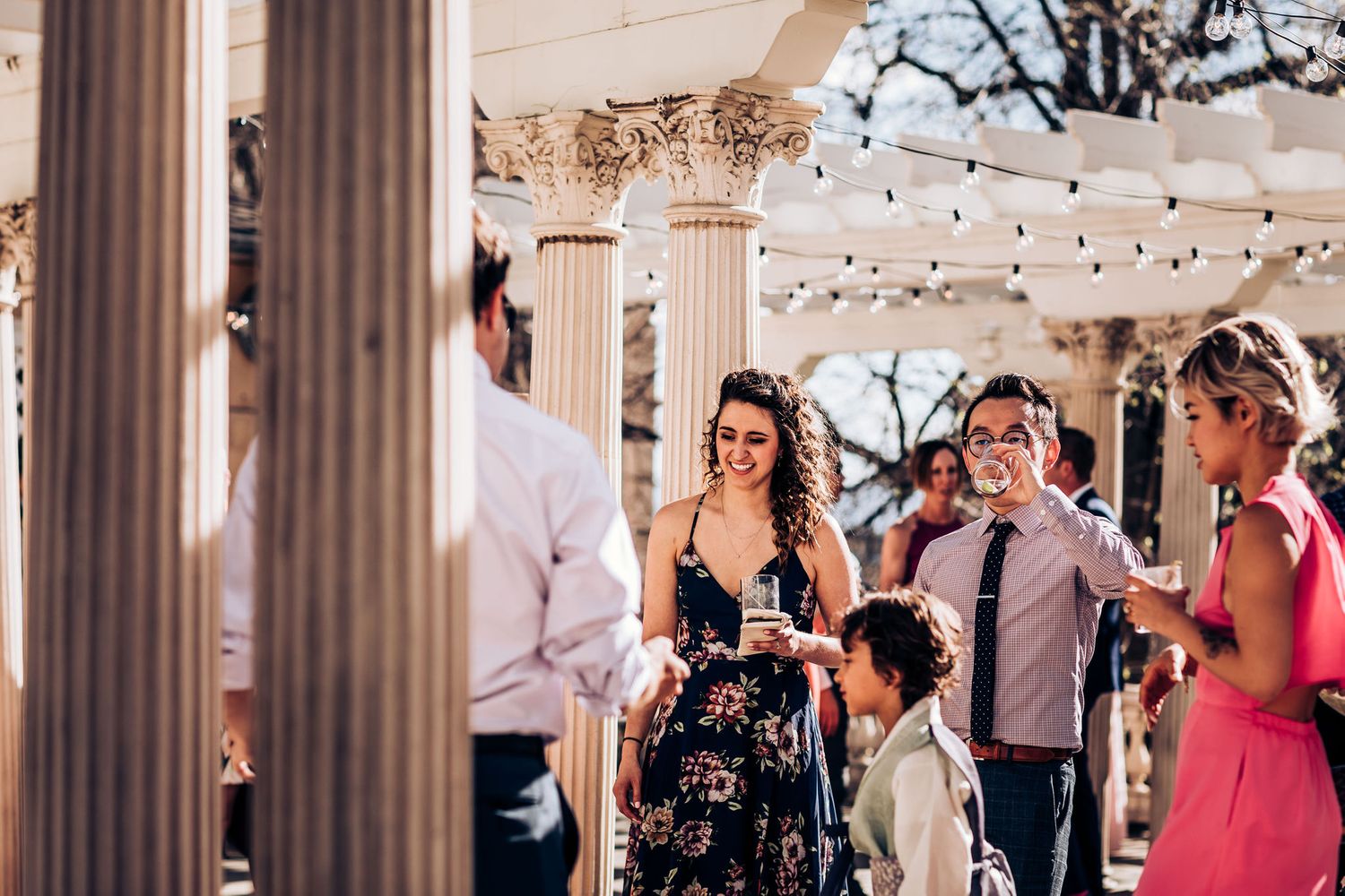 Guests mingle at an elegant outdoor reception with string lights and classical columns.