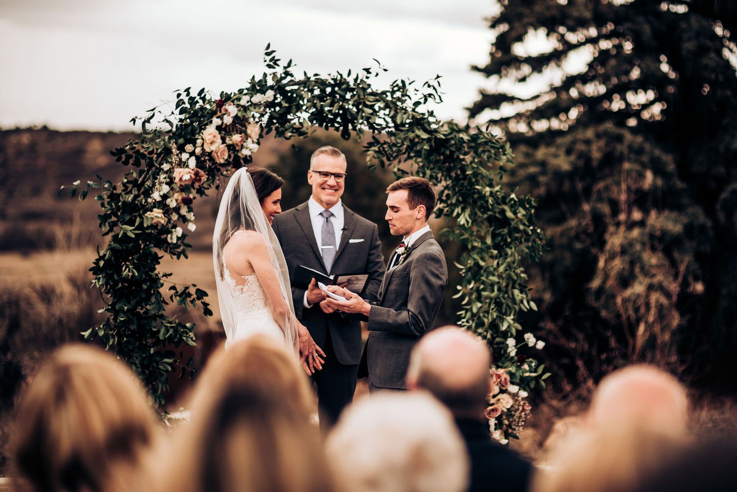 Outdoor wedding ceremony under a lush greenery arch with gathered guests and mountains in background.