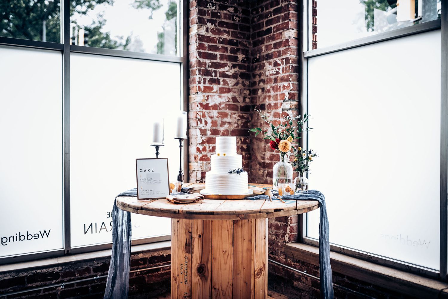 White wedding cake displayed on a rustic wooden table against exposed brick wall and large windows.