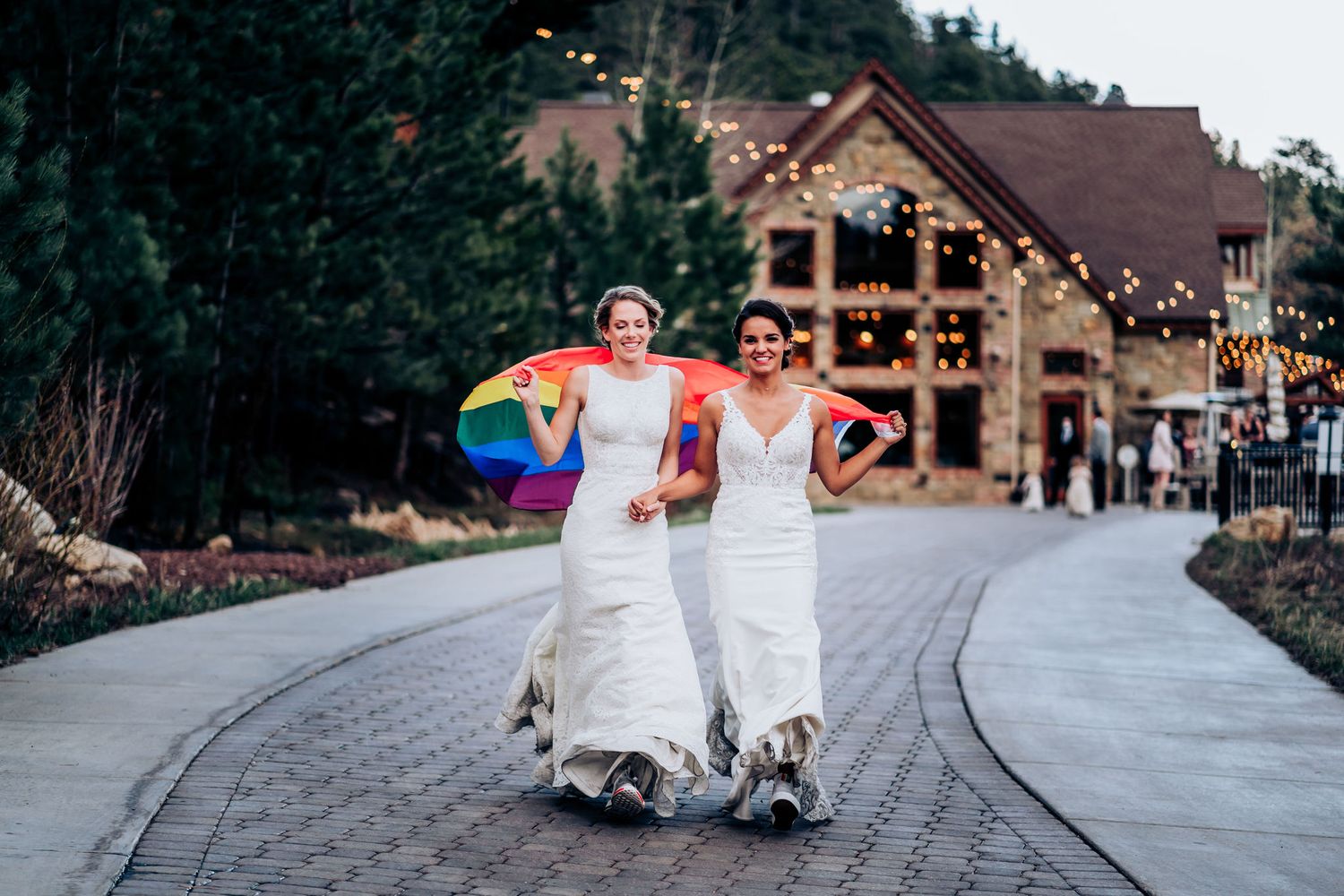 Two people in white wedding dresses walk together on a cobblestone path holding a rainbow flag with a rustic building behind them.