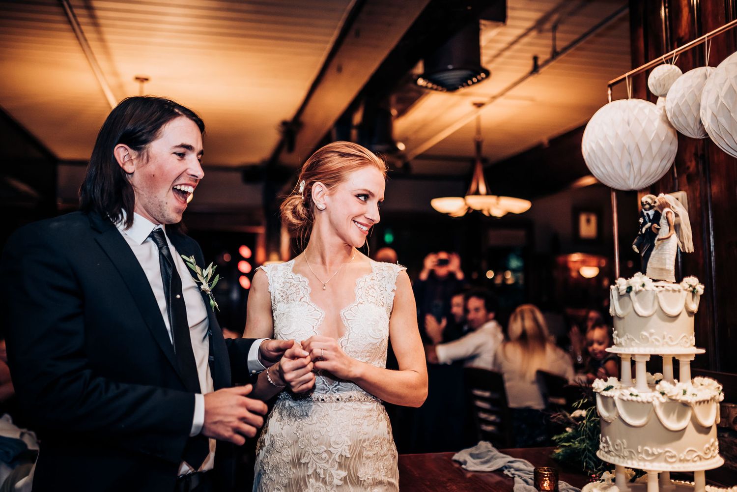 Couple stands beside their elegant white wedding cake decorated with paper lanterns in the background.