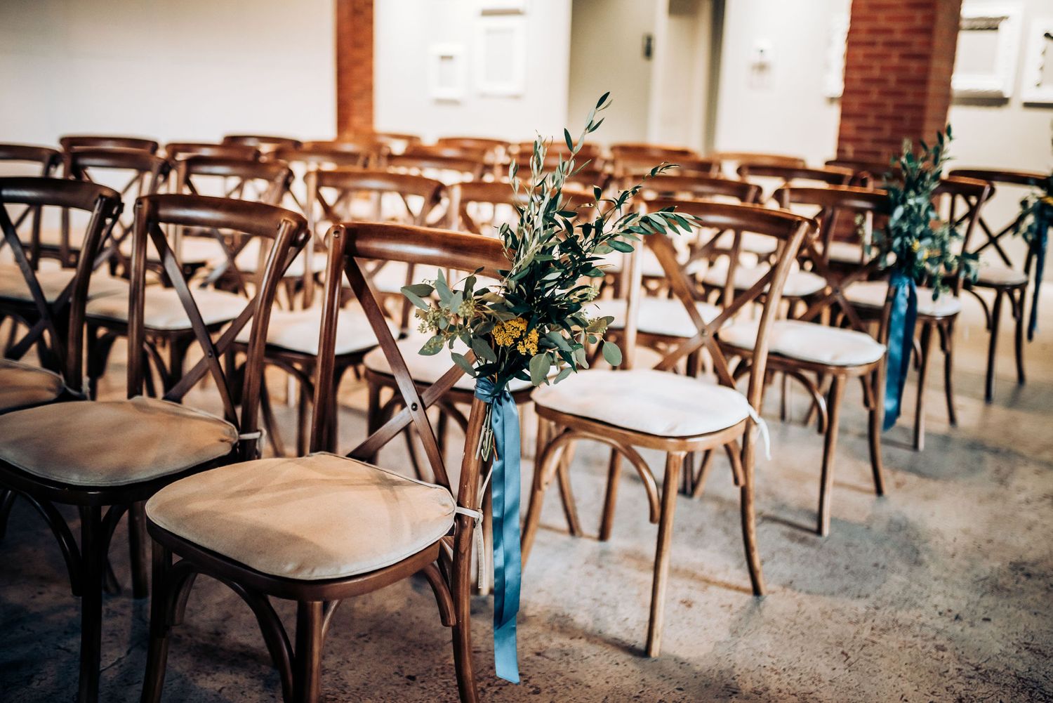 Rustic wedding ceremony setup with wooden chairs decorated with blue ribbons and floral arrangements.