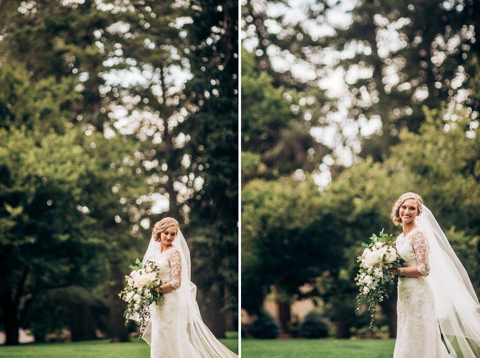 Bride in lace gown holds cascading bouquet while posing against green outdoor backdrop.