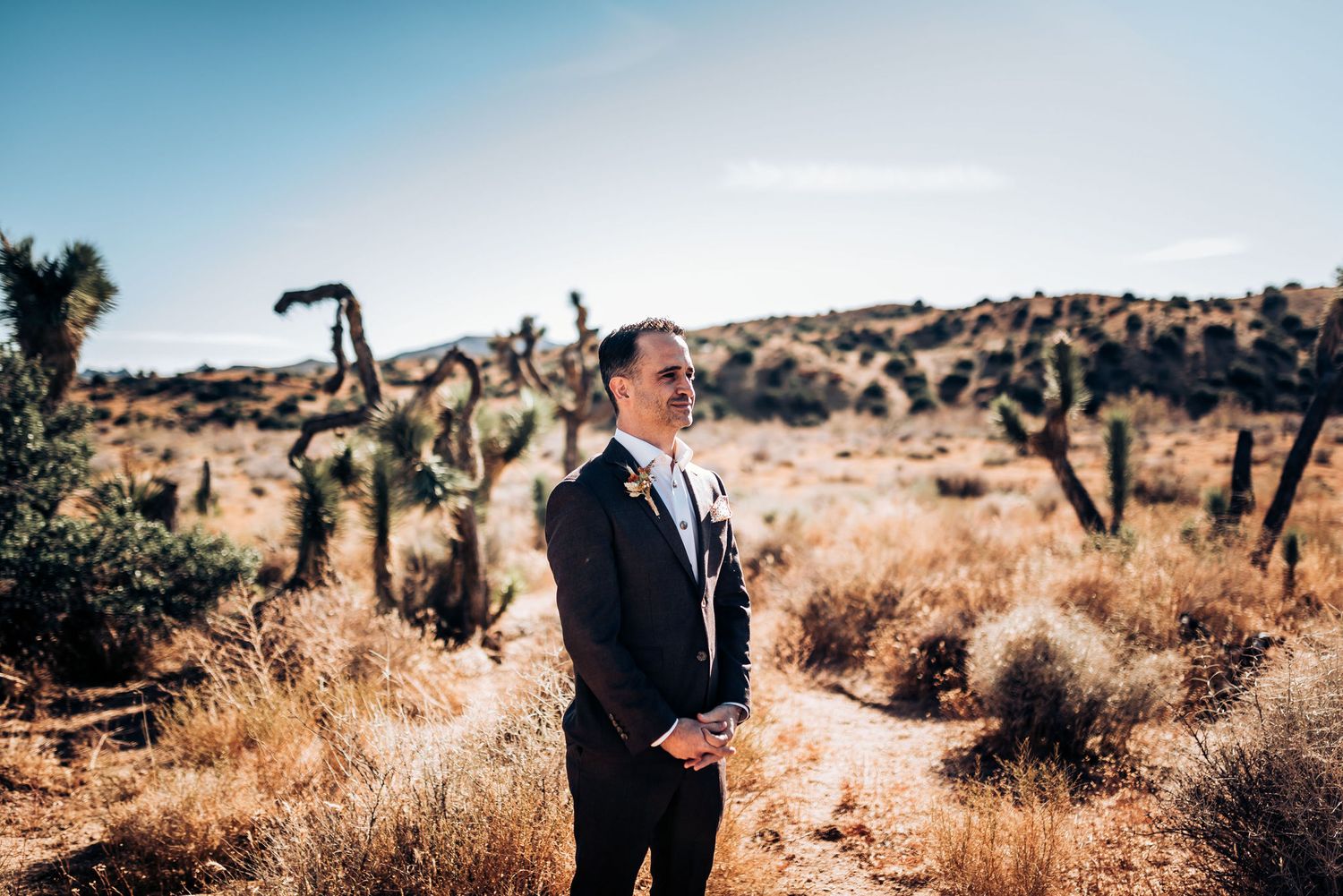 Groom stands in desert landscape surrounded by Joshua trees under blue sky.